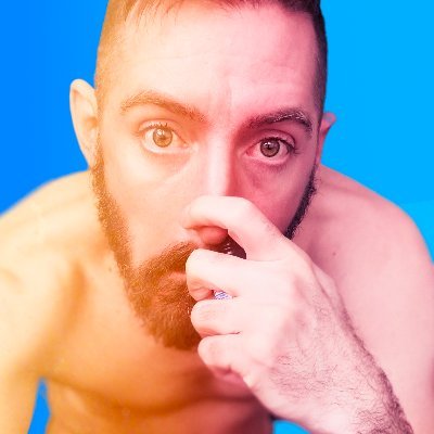 A close-up portrait of a shirtless man with a beard, holding his hand near his face, set against a bright blue background with warm orange and pink lighting.