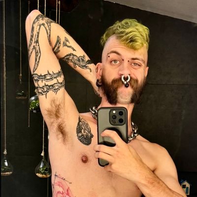 A shirtless man with tattoos, a green-tinted mohawk, and nose ring taking a mirror selfie with a black smartphone in a dark room.