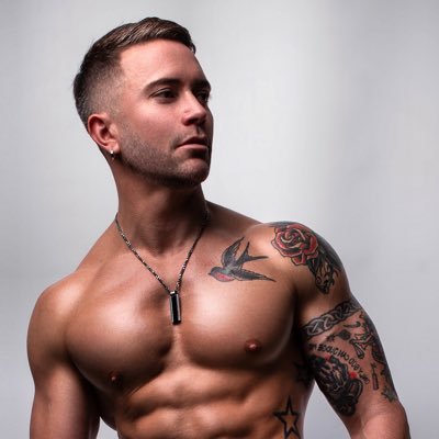A muscular man with tattoos and a beard, wearing a black pendant necklace, shown from the chest up against a light gray studio background.